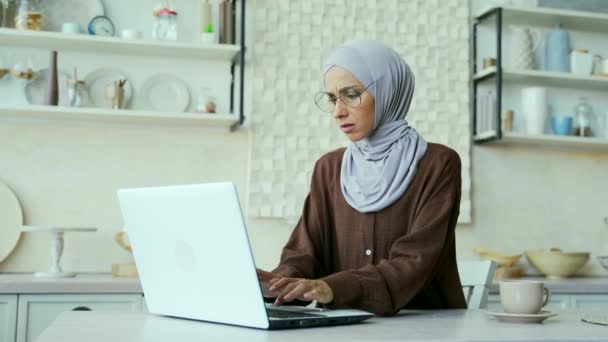 眼鏡をかけたヒジャーブのイスラム教徒の女性は 台所に座っているラップトップの問題を抱えていらいらしていますシステムエラー データ損失 バックアップバッテリーの問題 自宅でパスワードを忘れたため 動揺した女の子は動作できません — ストック動画