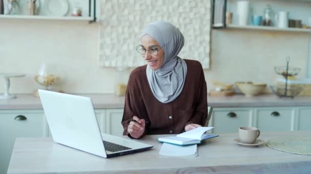ヒジャーブ州の若いムスリム女性を笑顔にし 眼鏡をかけ遠隔授業を受ける教師がノートを作り 室内のラップトップ画面を見る遠隔オンライン教育の概念 — ストック動画