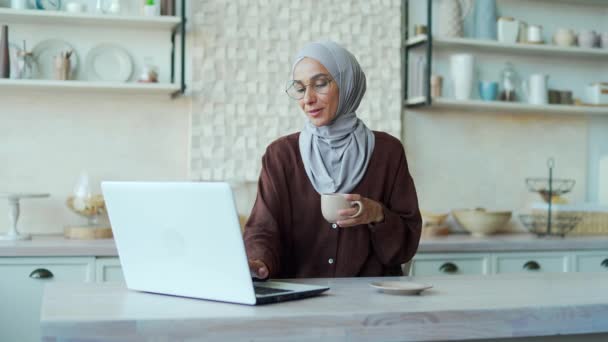 ヒジャーブ州でコーヒーや紅茶を楽しむムスリム女性の笑顔は 光の部屋でノートパソコンで彼女の仕事を続けます魅力的な女性は オンライン仕事や家庭から勉強ホームウォッチビデオでウェブを閲覧します — ストック動画