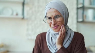 Stressed young Muslim woman with severe toothache dental pain ache using laptop computer at home alone Sad female suffering from discomfort because caries gum disease or sensitive teeth indoors