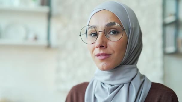 カメラを見て微笑むメガネ姿の若いイスラム教徒女性の頭部を撮影したポートレート 閉鎖顔の可愛いですArab女性でHihab屋内でホームチアリーディング女の子身に着けていますスカーフ閉じるアップ — ストック動画