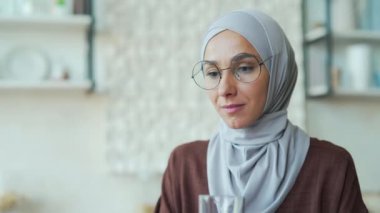 Muslim woman freelancer drinks clean water while sitting at table at home Happy female with a glass in her hands is quenching her thirst and enjoying herself indoor purified filtered mineral water