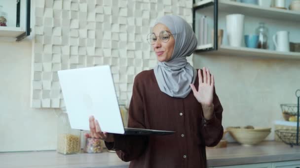 眼鏡をかけたヒジャーブのイスラム教徒の女性は ウェブカメラのラップトップコンピュータの挨拶を見て 同僚や学生にビデオ通話オンライン会議チャットで話します 家庭教師教育研究でリモートで — ストック動画