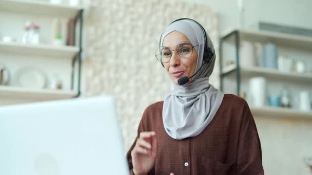 ヒジャーブの若いイスラム教徒の女性がビデオ通話で遠隔講義を行うビジネス女性教師や講師がオンライン会議や会議で話すラップトップコンピュータを使って自宅で座って勉強する — ストック動画