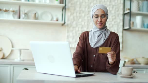 台所のラップトップコンピュータでクレジットカードで支払う眼鏡でヒジャーブの若いイスラム教徒の女性幸せな顧客は インターネットストアでオンラインショッピングをし 自宅で現金を受け取る簡単に支払う — ストック動画