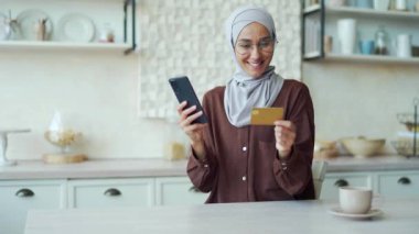 Young Muslim woman in hijab with glasses paying with credit card on smartphone in the kitchen Happy customer doing payments online shopping in internet store and receiving cashback indoors Easy pay