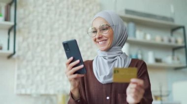 Young Muslim woman in hijab with glasses paying with credit card on smartphone in the kitchen Happy customer doing payments online shopping in internet store and receiving cashback indoors Easy pay