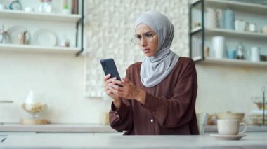 Scared young muslim woman in hijab read bad news browsing social media in smartphone while sitting at home Upset Confused female stares at screen and looks puzzled, unexpected problem fear