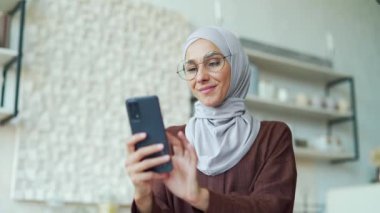 Gözlüklü gülümseyen esmer, akıllı telefonu tutuyor ve açık yerli mutfağında sosyal medyayı izliyor. Genç, mutlu, Arap kadın internet ürünleri çevrimiçi internet mağazasında kapalı mekanda telefonla geziniyor.