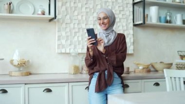 Gözlüklü gülümseyen esmer, akıllı telefonu tutuyor ve açık yerli mutfağında sosyal medyayı izliyor. Genç, mutlu, Arap kadın internet ürünleri çevrimiçi internet mağazasında kapalı mekanda telefonla geziniyor.
