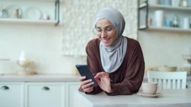 Gözlüklü gülümseyen esmer, akıllı telefonu tutuyor ve açık yerli mutfağında sosyal medyayı izliyor. Genç, mutlu, Arap kadın internet ürünleri çevrimiçi internet mağazasında kapalı mekanda telefonla geziniyor.