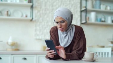 Scared young muslim woman in hijab read bad news browsing social media in smartphone while sitting at home Upset Confused female stares at screen and looks puzzled, unexpected problem fear