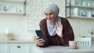 Genç müslüman kadın internette sohbet ediyor. Evde, mutfakta otururken uzaktan cep telefonu kullanıyor. Tesettürlü gülümseyen kadın akıllı telefondan arkadaşlarıyla konuşuyor. Hücre kamerasını kullan.