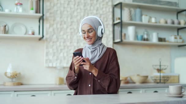 ヘッドフォンでヒジャーブの魅力的なイスラム教徒の女性は 彼女のスマートフォンで歌を選択し 軽いキッチンで踊ります音楽を聞いて眼鏡をかけて若い女性を笑顔と携帯電話を使用して自宅で楽しみを持っています — ストック動画
