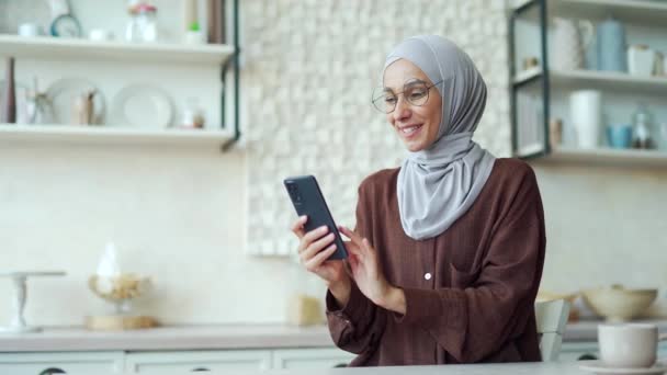 スマートフォンのスクロールや台所でソーシャルメディアを見てメガネを持つヒジャーブの女の子の笑顔イスラム教徒の女性自宅の屋内で携帯電話のショッピングオンラインストアを使用して若い女性 — ストック動画