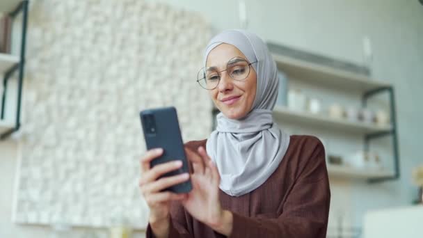 スマートフォンのスクロールを保持し 軽量家庭用キッチンでソーシャルメディアを見てメガネとスマイリングブルネット若い幸せなアラブ女性は屋内で電話でオンラインインターネットストアで製品を閲覧 — ストック動画