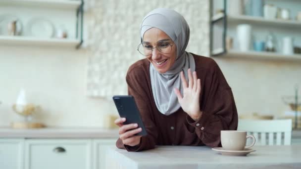 若いイスラム教徒の女性は台所に座っている間に携帯電話を使って遠隔でビデオ通話でオンラインでチャットします 笑顔女性でヒジャーブ語で友人と話す上のスマートフォン挨拶使用細胞ウェブカメラ — ストック動画