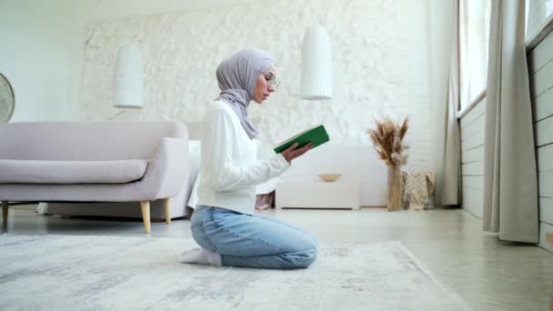 ヒジャーブの若いムスリム女性が自宅で祈ります ラウンジでベールを身に着けている間 クルアーンを読む神への崇拝家の明るいリビングルームで一人で東文化宗教ライフスタイル — ストック動画