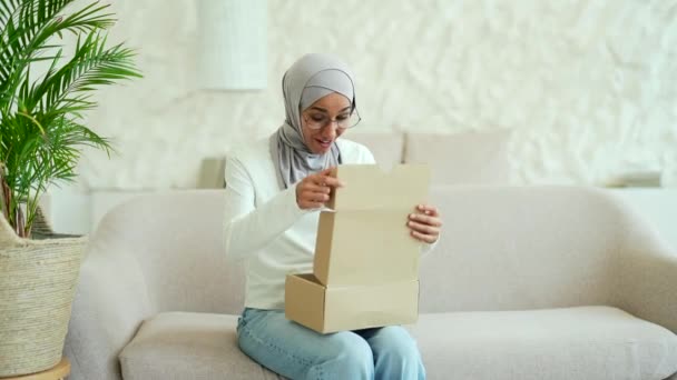 ヒジャーブ州の若いイスラム教徒の女性は自宅でソファに座っているオンラインインターネットストアでの買い物に満足した小包を受け取りましたカートンボックスを屋内で開きながら幸せな顧客は購入注文で喜びます — ストック動画