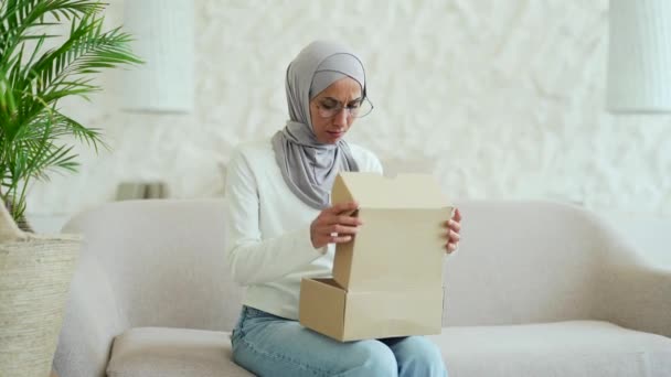 ヒジャーブ州の若いイスラム教徒の女性は自宅でソファに座っているオンラインインターネットストアでの買い物に不満を小包を受け取りました室内でカートンボックスを開きながら 顧客は購入注文に失望しました — ストック動画