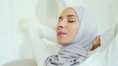 Close up of beautiful young Muslim woman in hijab relaxing on comfortable sofa with hands behind head in the light living room Smiling peaceful girl enjoy calm time at home alone No stress concept