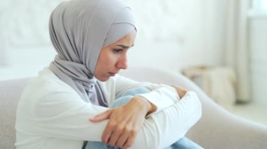 Sad depressed Muslim woman in hijab feels unhappy by psychological problem break up or hard divorce Mental health thinking of personal problems alone at home worried suffering girl sitting on the sofa