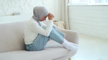 Sad depressed Muslim woman in hijab feels unhappy by psychological problem break up or hard divorce Mental health thinking of personal problems alone at home worried suffering girl sitting on the sofa