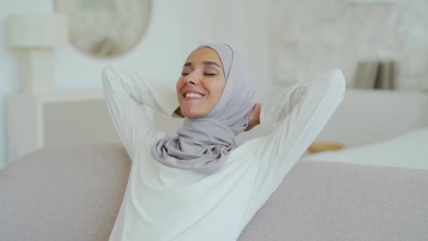 美しい若いイスラム教徒の女性の閉じ込めますヒジャーブで快適なソファの上で光のリビングルームで手を後ろにリラックス笑顔平和的な女の子は家だけで穏やかな時間をお楽しみくださいストレスコンセプトなし — ストック動画