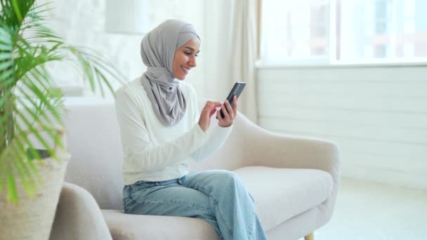 ヒジャーブ州のムスリム女性の笑顔自宅のリビングルームでスマートフォンのスクロールやソーシャルメディアを見て携帯電話のテキストメッセージを使用してオンラインストアで若い女性のブラウジング製品屋内でチャット — ストック動画