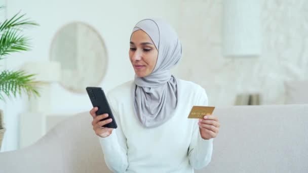 自宅でスマートフォンの携帯電話でクレジットカードで支払うメガネを持つヒジャーブの若いイスラム教徒の女性幸せな顧客は 屋内で現金を受け取るインターネットストアでオンラインショッピングを行う簡単に支払う — ストック動画