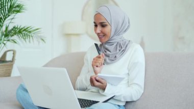 Smiling young muslim woman in hijab student having a remote lesson listening teacher lecturer making notes video course and looking at laptop screen in living room indoor Distance online education