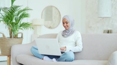 Smiling Muslim girl in hijab freelancer working on laptop checking email surfing web typing at living room workplace indoors young woman student has online distance education on computer at home