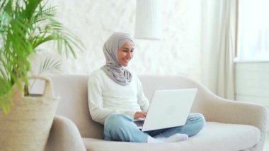 Smiling Muslim girl in hijab freelancer working on laptop checking email surfing web typing at living room workplace indoors young woman student has online distance education on computer at home