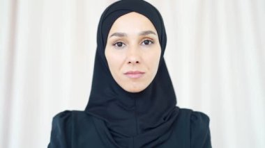 closeup portrait of a pretty young Muslim woman in a black hijab standing and smiling indoors at home Headshot of an Islamic happy female looking at the camera on a light room background close up