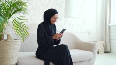 Smiling Muslim woman in hijab hold smartphone scrolling and watching social media in living room at home Young female browsing products in online internet store using phone texting chatting indoors