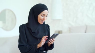 Smiling Muslim woman in hijab hold smartphone scrolling and watching social media in living room at home Young female browsing products in online internet store using phone texting chatting indoors