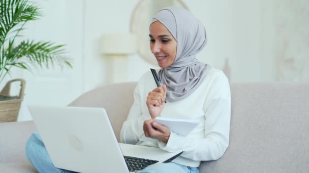 ヒジャーブ州の若いムスリム女性が遠隔授業を受けている講師がノートビデオコースを作成し リビングルームの屋内でラップトップ画面を見る遠隔オンライン教育 — ストック動画
