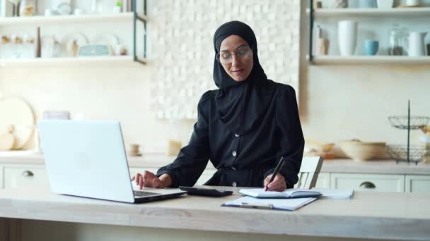 若いイスラム教徒の女性は オンラインで家庭から仕事を計算したり リモートで勉強しますヒジャーブの女性はノートパソコンを使用してコンピュータノートビジネス女性は アプリやウェブサイトを使用して個人的な財政を管理します — ストック動画