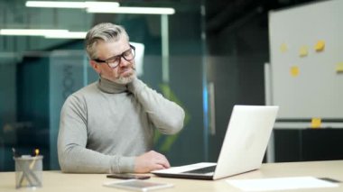 Mature gray haired bearded businessman wearing glasses has neck pain while sitting at desk at workplace in modern office. A tired middle aged entrepreneur rubs and massages his muscles with his hands