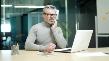 Mature gray haired bearded businessman wearing glasses has a toothache while sitting at a desk at a workplace in modern office. Tired middle aged entrepreneur holding hand on cheek massaging sore spot