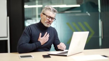 Sick mature gray haired bearded businessman in glasses has heartache sitting at desk at workplace in modern office. Tired middle aged entrepreneur having a heart attack, holding his hand on his chest