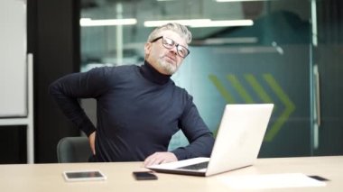 Sick mature gray haired bearded businessman in glasses has back pain while sitting at desk at workplace in modern office. Tired middle aged man holds a hand on his lower back and massages a sore spot
