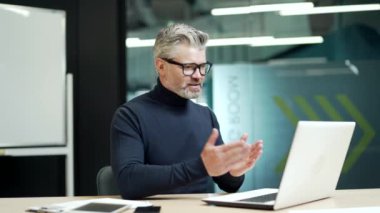 Modern mature gray haired bearded man is making a video call using a laptop while sitting at a desk at a workplace in a modern office. A confident entrepreneur shares his ideas with a business partner