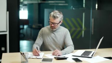 Mature gray haired bearded businessman in glasses is calculating on a calculator and writing in a notebook while sitting at a desk in office. Entrepreneur calculates the costs of the business project