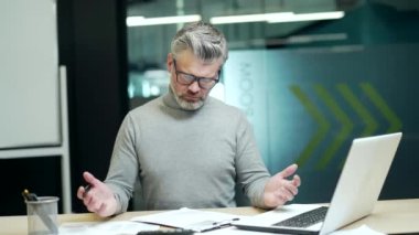 Confused mature gray haired bearded businessman in glasses having difficulty with paper work while sitting at desk in office. A dissatisfied entrepreneur fills out a document he does not understand