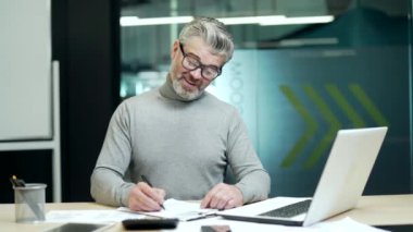 Smiling mature gray haired bearded businessman in glasses making notes in notebook at desk at workplace in modern office. Happy entrepreneur owner making plans for the day showing hand gesture yes