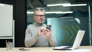 Serious mature gray haired bearded businessman in glasses carefully reading message on smartphone at workplace in modern office. A concentrated entrepreneur is thinking about an important problem