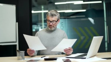 Smiling mature gray haired bearded businessman looking through business papers at desk at workplace in modern office. The happy entrepreneur is satisfied with what he has read and signs the documents