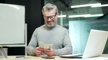 Cheerful mature gray haired bearded businessman in glasses counts dollar bills at desk at workplace in modern office. A happy entrepreneur rejoices at good earnings and holds money in his hands