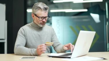 Sad mature gray haired bearded businessman having problem typing credit card number on laptop at workplace in office. Entrepreneur is angry because he faced fraud, money was stolen from his account